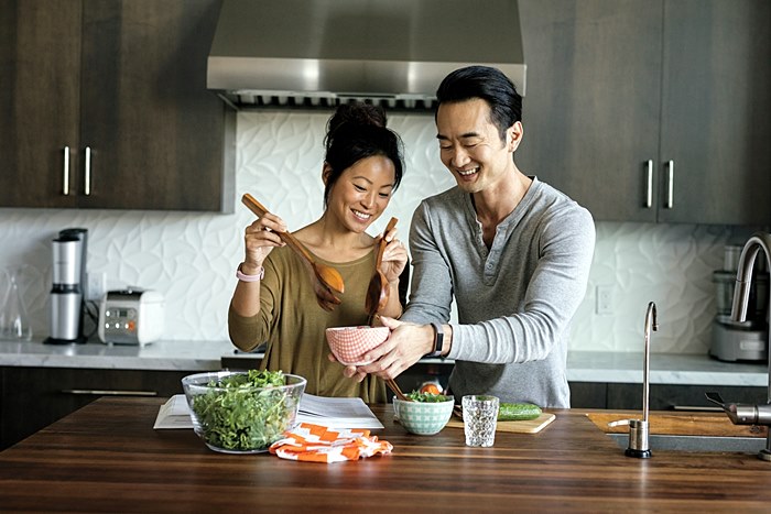 Alta HR Lifestyle photo featuring a young asian couple cooking with the woman wearing a special edition tracker in rose gold and soft pink, the man wearing a tracker with a blue gray band.